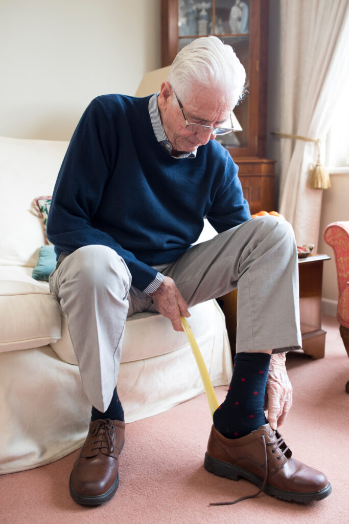 Ein älterer Herr mit weißen Haaren sitzt auf einem hellen Sofa und nutzt einen Schuhanzieher, um seine braunen Lederschuhe anzuziehen.