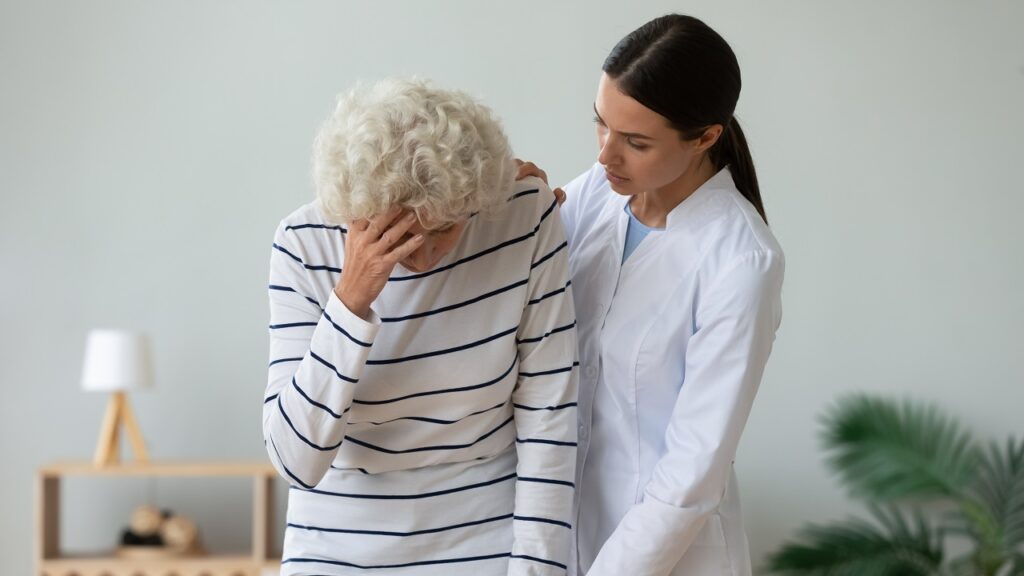 Eine Seniorin in gestreiften Shirt hält sich den Kopf, während eine Pflegerin ihr beruhigend die Hand auf die linke Schulter legt.